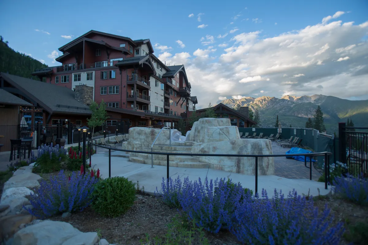 A tranquil exterior view featuring a stone fountain with colorful flowers in bloom, set against a dramatic mountain backdrop and the resort building. There are outdoor seating areas ideal for gathering, relaxing, and enjoying the scenery together.