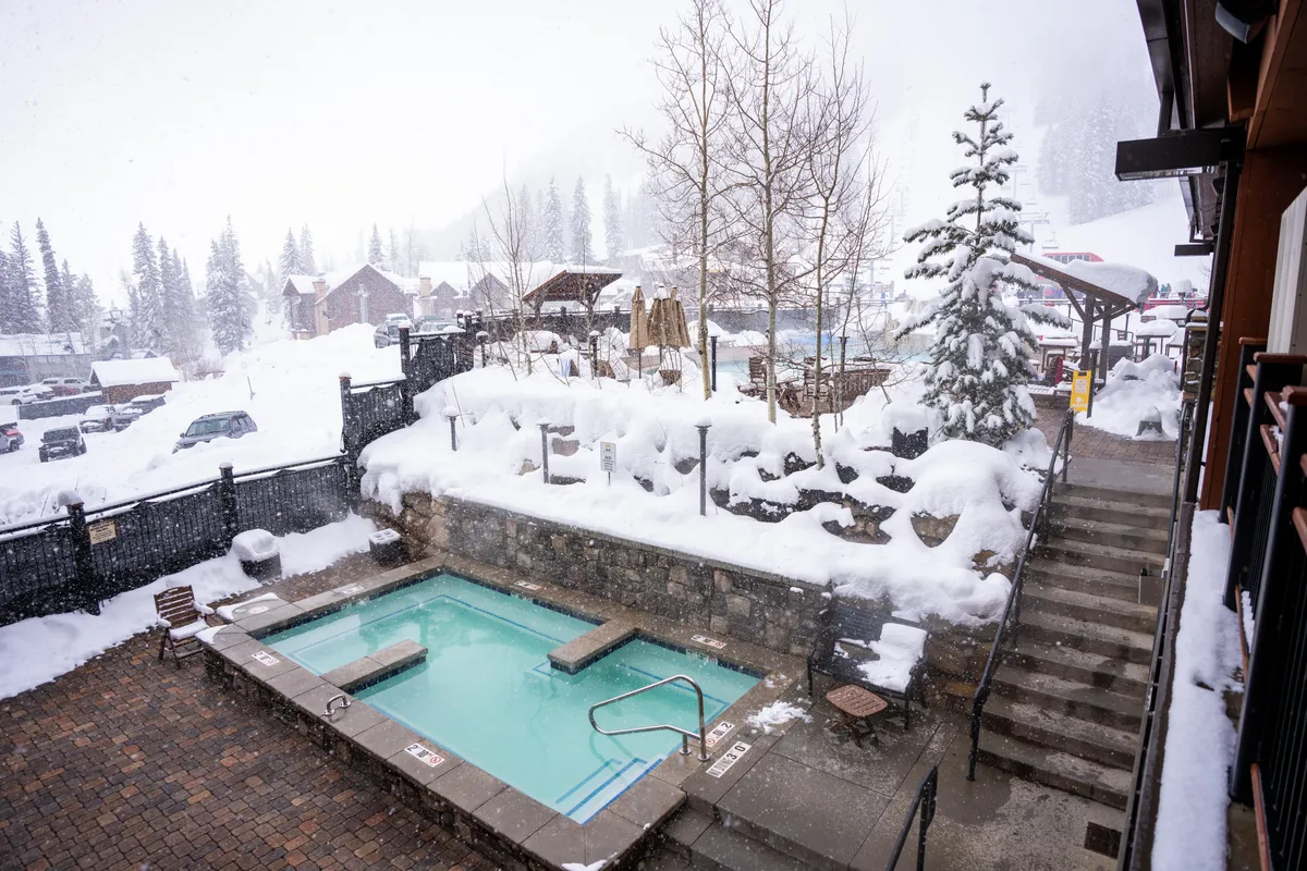 A tranquil exterior shot featuring a steaming hot tub surrounded by snow-covered pine trees and stone walls. The cozy setting invites relaxation amid a wintry landscape, perfect for unwinding after a day of adventure.