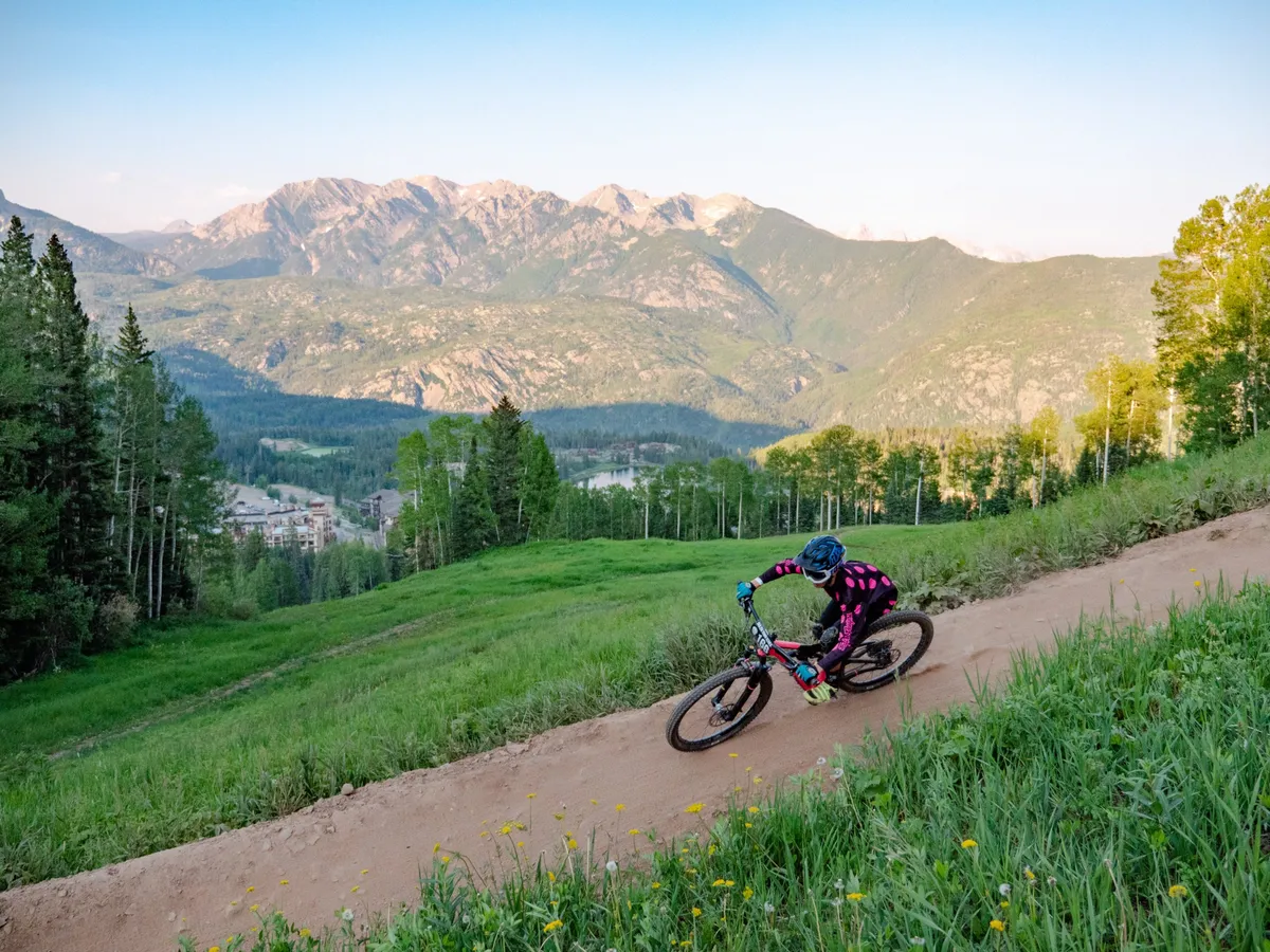 An energetic exterior scene showcasing a mountain biker riding along a winding trail with lush greenery and majestic mountain peaks in the background. The vibrant landscape suggests a variety of outdoor activities and captures the adventurous spirit of the San Juan area.