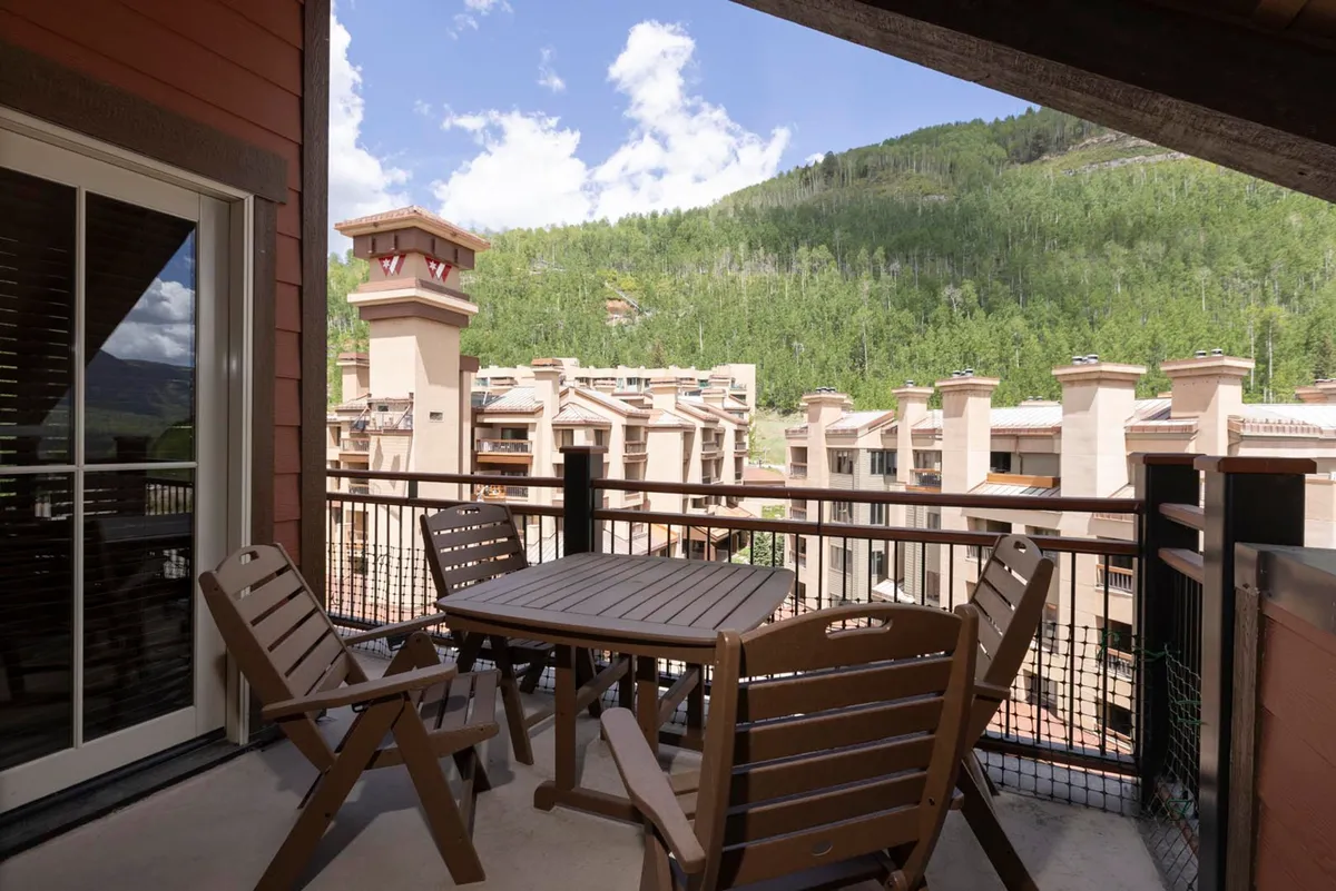 A private outdoor balcony with a rustic wooden table and chairs overlooks a sweeping mountain landscape under a blue sky. The space invites guests to dine al fresco, sip morning coffee, or toast the sunset with friends, while taking in the tranquil natural surroundings.