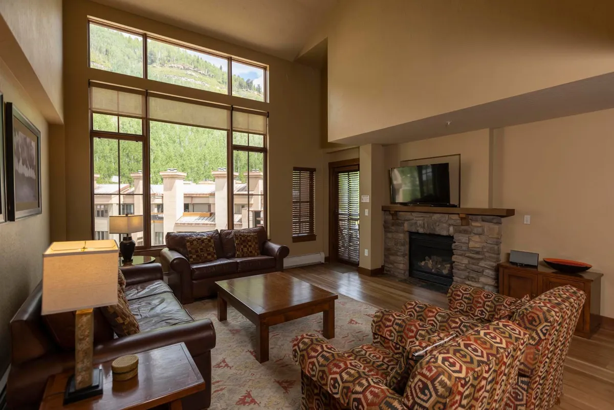 The image showcases a spacious living area with plush leather sofas and colorful patterned chairs arranged around a striking stone fireplace. Floor-to-ceiling windows frame sweeping views of snow-capped mountains, bathing the room in natural light and offering a breathtaking backdrop. The cozy yet elegant space is perfect for both family relaxation and entertaining guests, embodying a warm mountain lodge atmosphere.