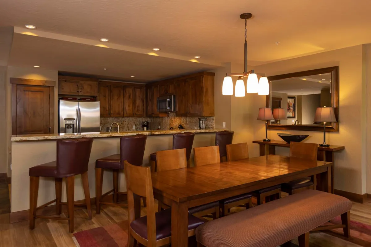 This image features a warm, inviting dining table set adjacent to a kitchen island, with stylish lighting fixtures above and rich wooden cabinetry throughout. The space is clearly designed for both everyday meals and special gatherings, with enough room to accommodate family and friends. The open layout naturally connects the kitchen and dining areas, fostering a communal atmosphere.