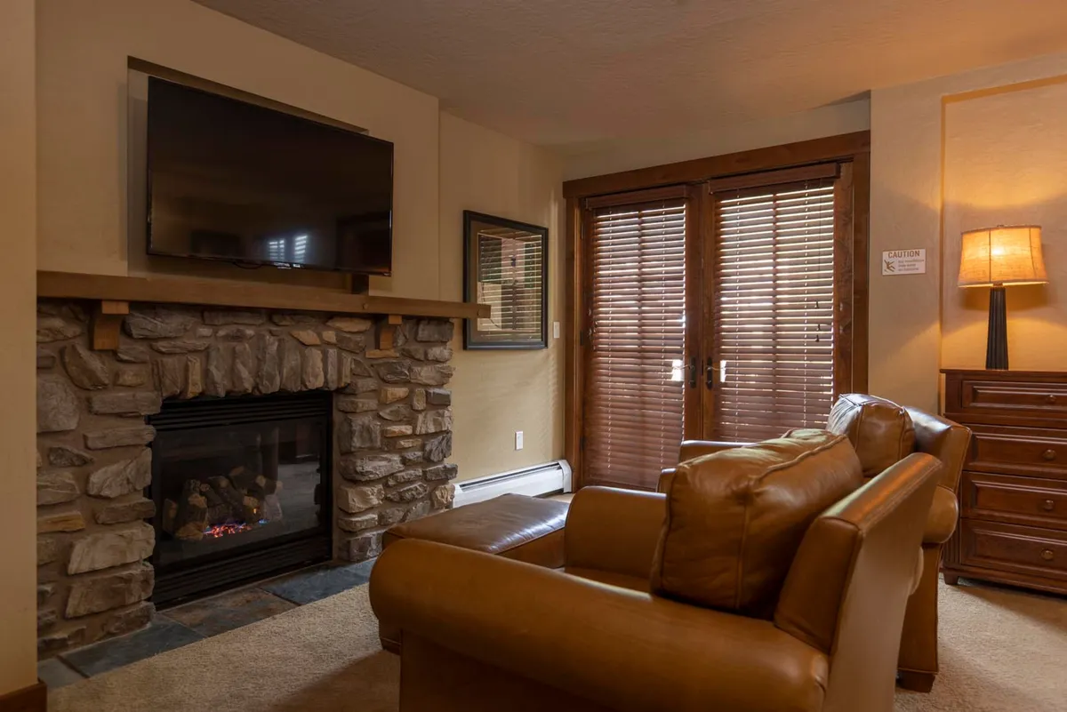 A warm living space featuring a striking stone fireplace as its focal point. The arrangement includes a leather armchair and wooden furniture, complemented by a TV for entertainment. The room is bathed in a soft, welcoming glow—ideal for relaxing by the fire or enjoying a cozy movie night after a day outdoors.