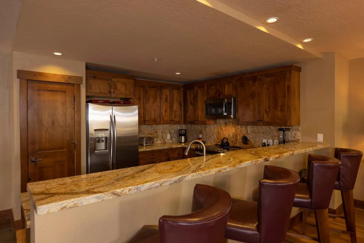 A closer view of the modern kitchen, this image spotlights the granite countertops, sleek stainless steel appliances, and comfortable bar stools, with wood cabinetry providing a touch of mountain lodge charm. The layout encourages socializing, whether preparing meals or enjoying a casual snack at the island.