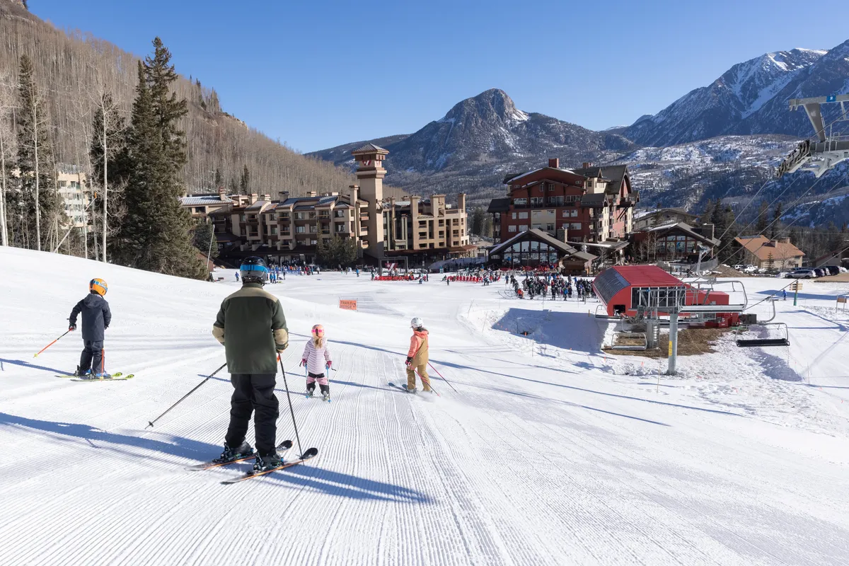 This vibrant exterior image features skiers and snowboarders actively enjoying the slopes of Purgatory Resort, with a dramatic mountain backdrop, resort buildings, and a ski lift in view. The lively scene captures families and individuals partaking in lessons and outdoor winter fun.