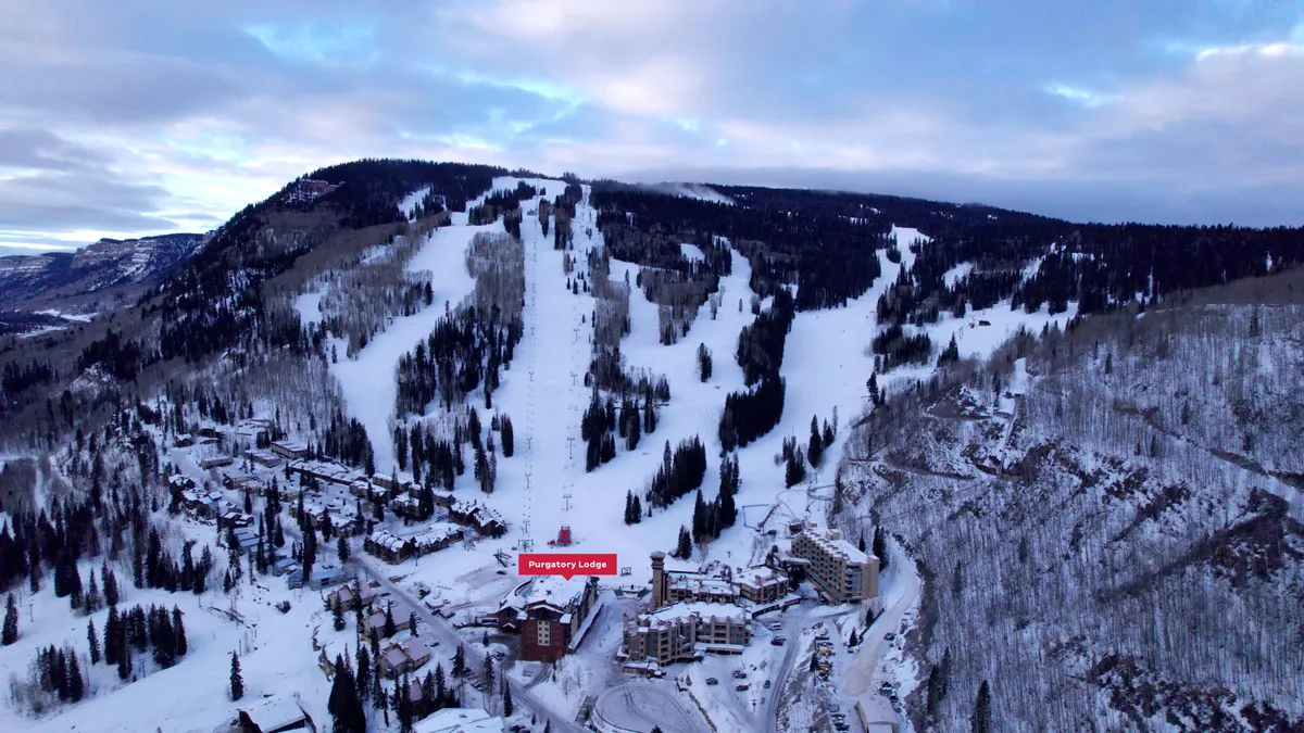 An exterior view of Purgatory Lodge nestled at the base of snow-covered ski slopes, framed by evergreen trees and majestic mountains. The scene includes guests relaxing around the lodge and hints of early morning or dusk light, evoking both adventure and coziness.